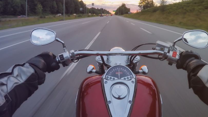 First-person view from a motorcycle traveling on a multi-lane highway at dusk, with the rider’s gloved hands on the handlebars and open road ahead