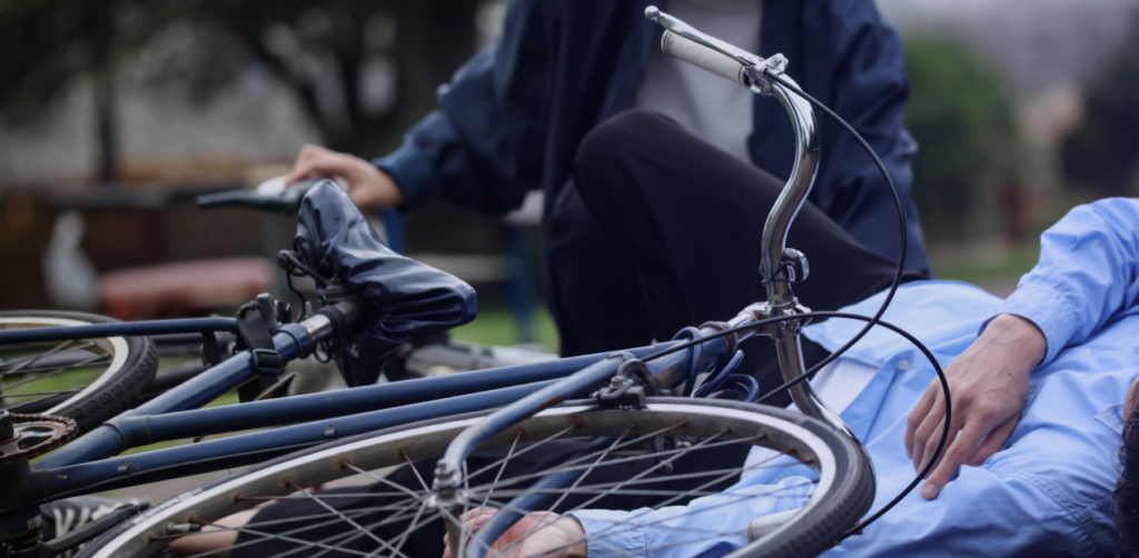 Injured cyclist lies on the ground beside a fallen bicycle while another person kneels nearby