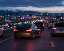 Heavy traffic fills a multilane city roadway at dusk, with glowing headlights and taillights beneath a mountain skyline