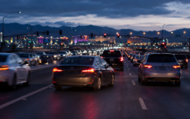 Heavy traffic fills a multilane city roadway at dusk, with glowing headlights and taillights beneath a mountain skyline