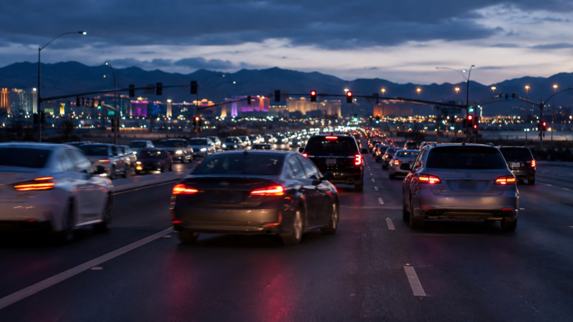 Heavy traffic fills a multilane city roadway at dusk, with glowing headlights and taillights beneath a mountain skyline