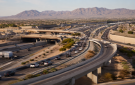 Wide aerial view of intersecting freeways, ramps, and flowing traffic surrounded by desert neighborhoods and mountains